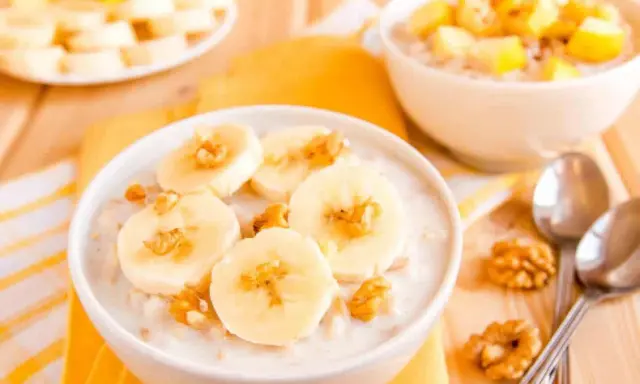 Un bol de talbina recette, garni de rondelles de banane et de noix. Une autre assiette de fruits est visible en arrière-plan.