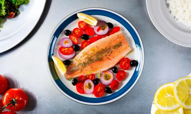 Une assiette colorée présente une truite au four, accompagnée de tomates cerises, d'oignons rouges, d'olives noires et de quartiers de citron.