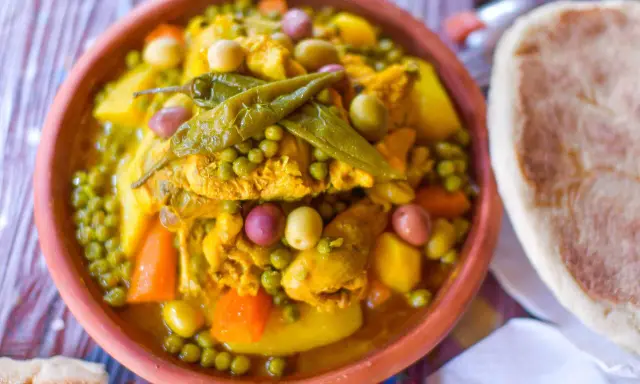 Un plat de tajine poulet aux olives, petits pois et carottes, garni de piments verts. Accompagné de pain.