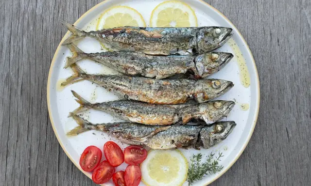 Cinq chinchards poissons grillés, servis sur une assiette avec des quartiers de citron, des tomates cerises et des herbes.
