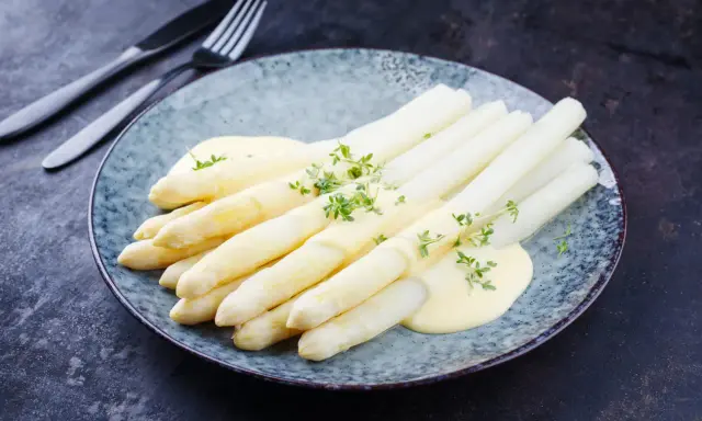 Asperges blanches nappées de sauce hollandaise et garnies d'herbes fraîches sur une assiette bleue.