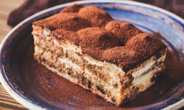 Tranche de gâteau tiramisu biscuit, généreusement saupoudrée de cacao, sur une assiette bleue.