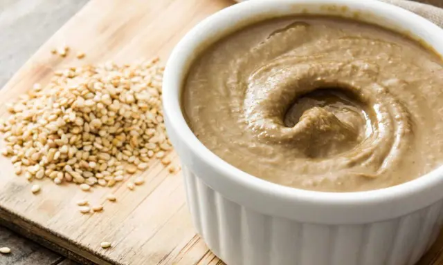 Un bol de tahini crémeux, prêt pour votre prochaine recette, à côté de graines de sésame sur une planche en bois.