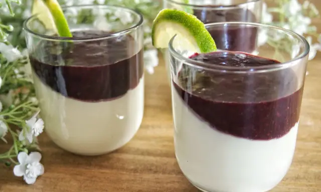 Trois panna cotta crémeuses, garnies d'un coulis pour panna cotta aux fruits rouges et d'une tranche de citron vert, sur un fond de fleurs blanches.