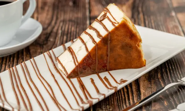 Tranche de san sebastian cheesecake, généreusement arrosée de sauce chocolat, sur une assiette blanche, avec une tasse de café en arrière-plan.
