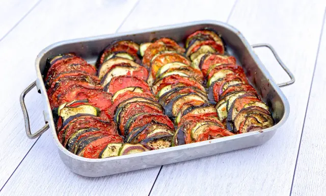 Un plat de tian de légumes, joliment arrangé en rondelles alternées de tomates, courgettes et aubergines, prêt à être dégusté.