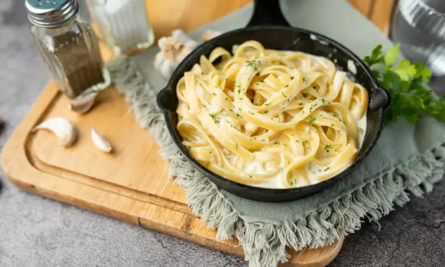 Un plat de fettuccine alfredo crémeux, garni de persil frais, servi dans une poêle en fonte. Des gousses d'ail et des moulins à sel et poivre sont disposés sur une planche à découper en bois.