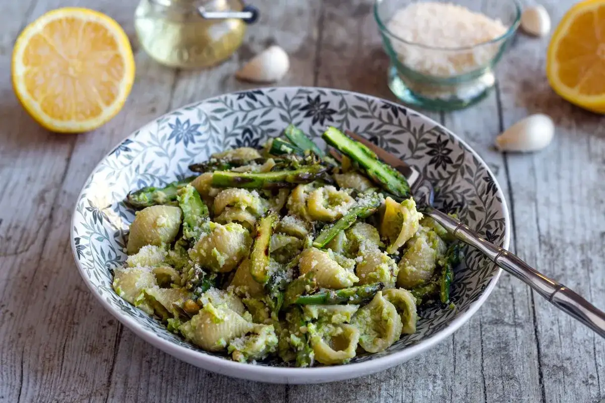 Un plat de pâtes aux asperges, saupoudré de parmesan, avec des citrons et de l'ail en arrière-plan.
