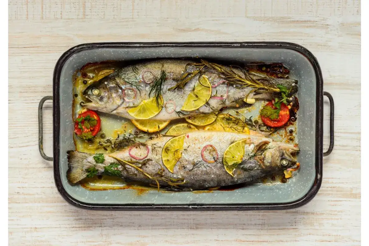Deux truites au four, garnies de rondelles de citron, d'oignon rouge, de tomates cerises et d'herbes aromatiques, dans un plat &eacute;maill&eacute;.