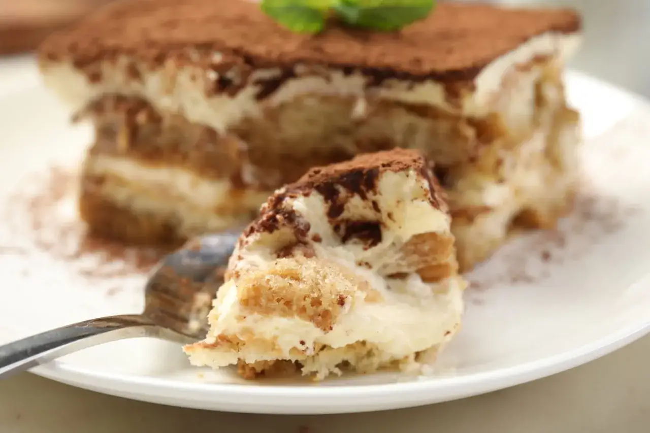 Une part de tiramisu, saupoudrée de cacao, attend sur une assiette. Une cuillère est prête à déguster ce délice.