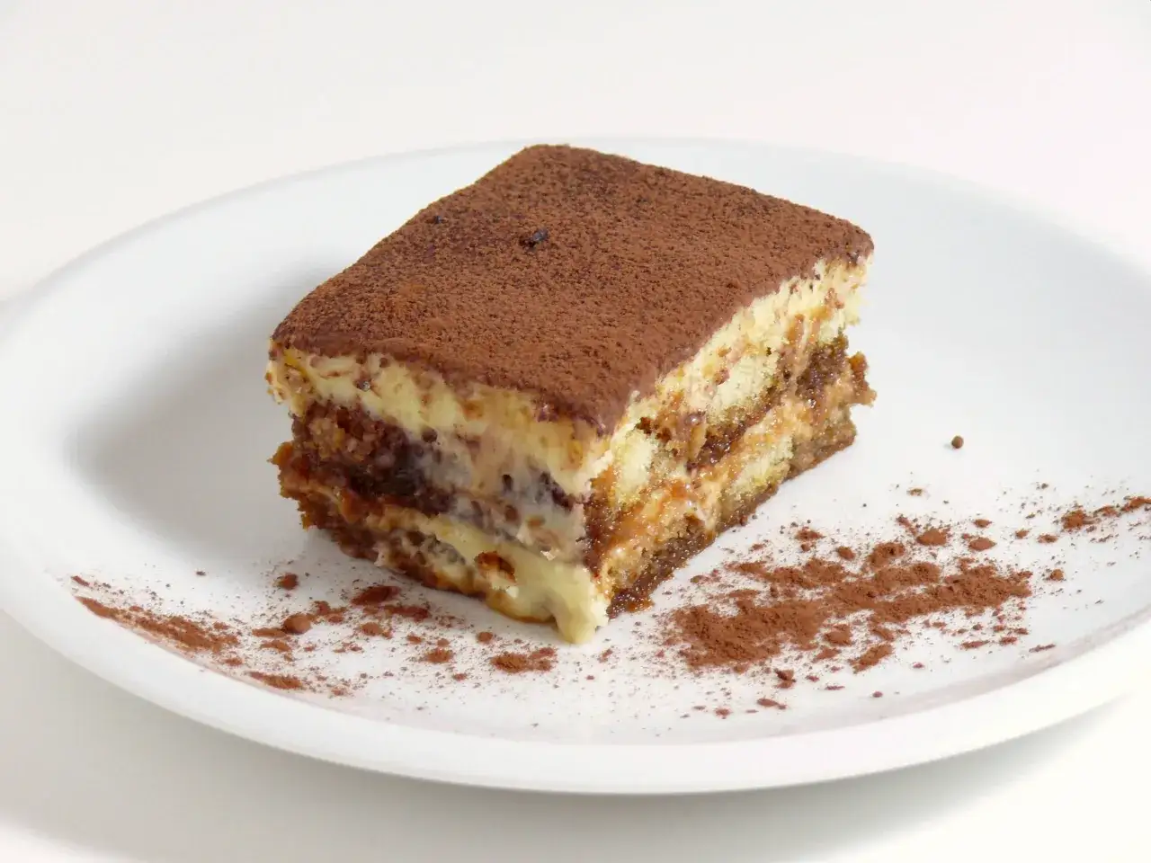 Une part de tiramisu, dessert aux origines italiennes, saupoudrée de cacao sur une assiette blanche.