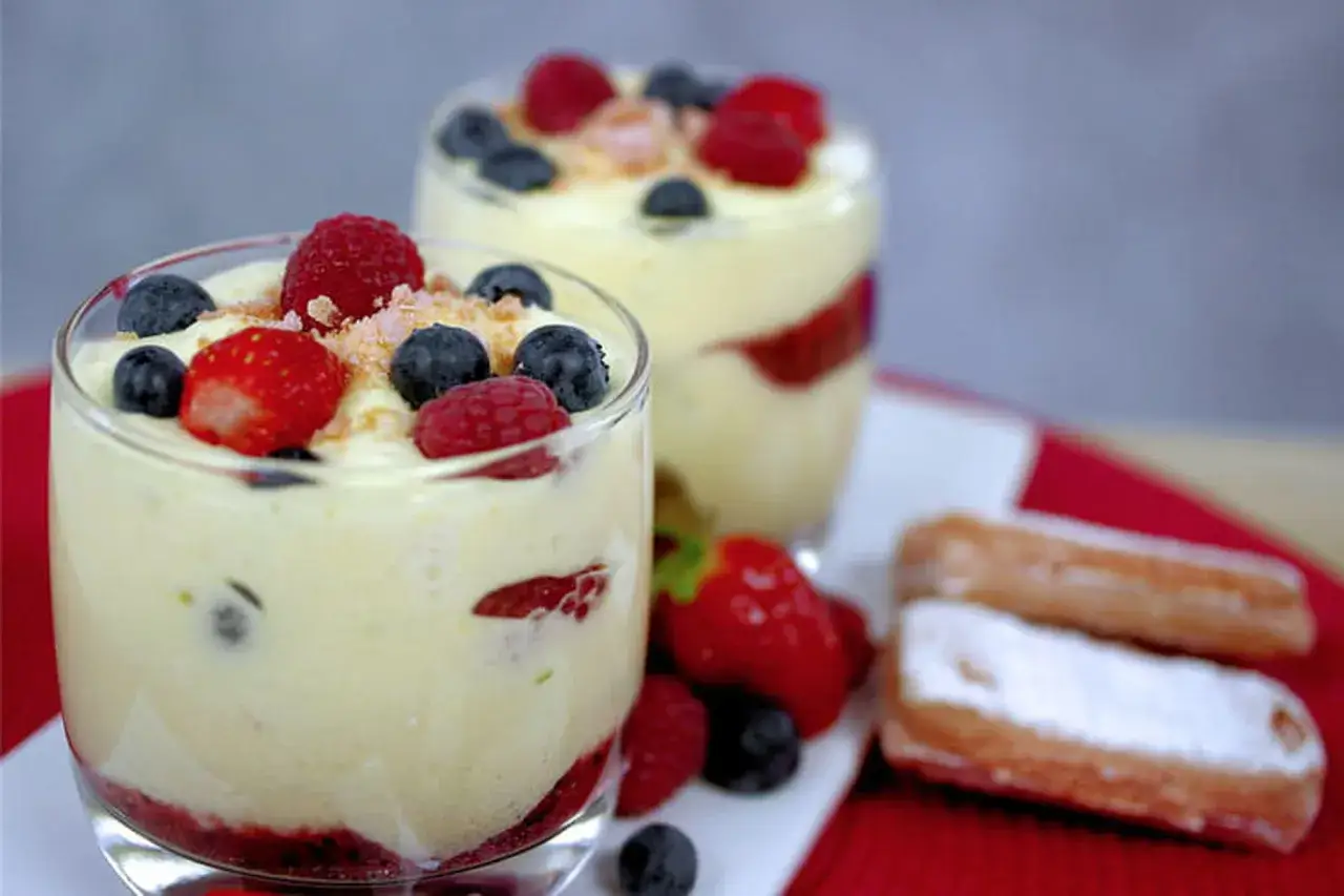 Deux verrines de tiramisu fruits rouges, garnies de framboises, myrtilles et fraises. Des biscuits à la cuillère saupoudrés de sucre glace sont à côté.