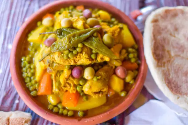 Un plat de tajine poulet aux olives, petits pois et carottes, garni de piments verts. Accompagné de pain.